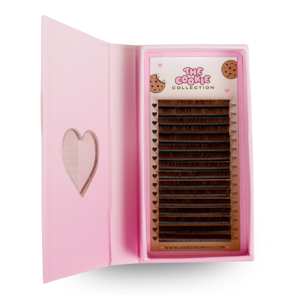 A pink presentation box for 'The Cookie Collection' eyelash extensions, shown open on a white background. The box features a heart-shaped cutout on the left panel and contains multiple rows of dark brown eyelash extensions on a pink tray.